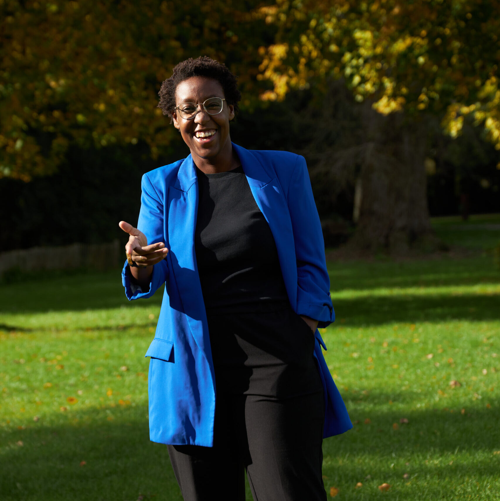 Belinda in a black shirt with a blue blazer smiling at the camera, against an autumn forest background